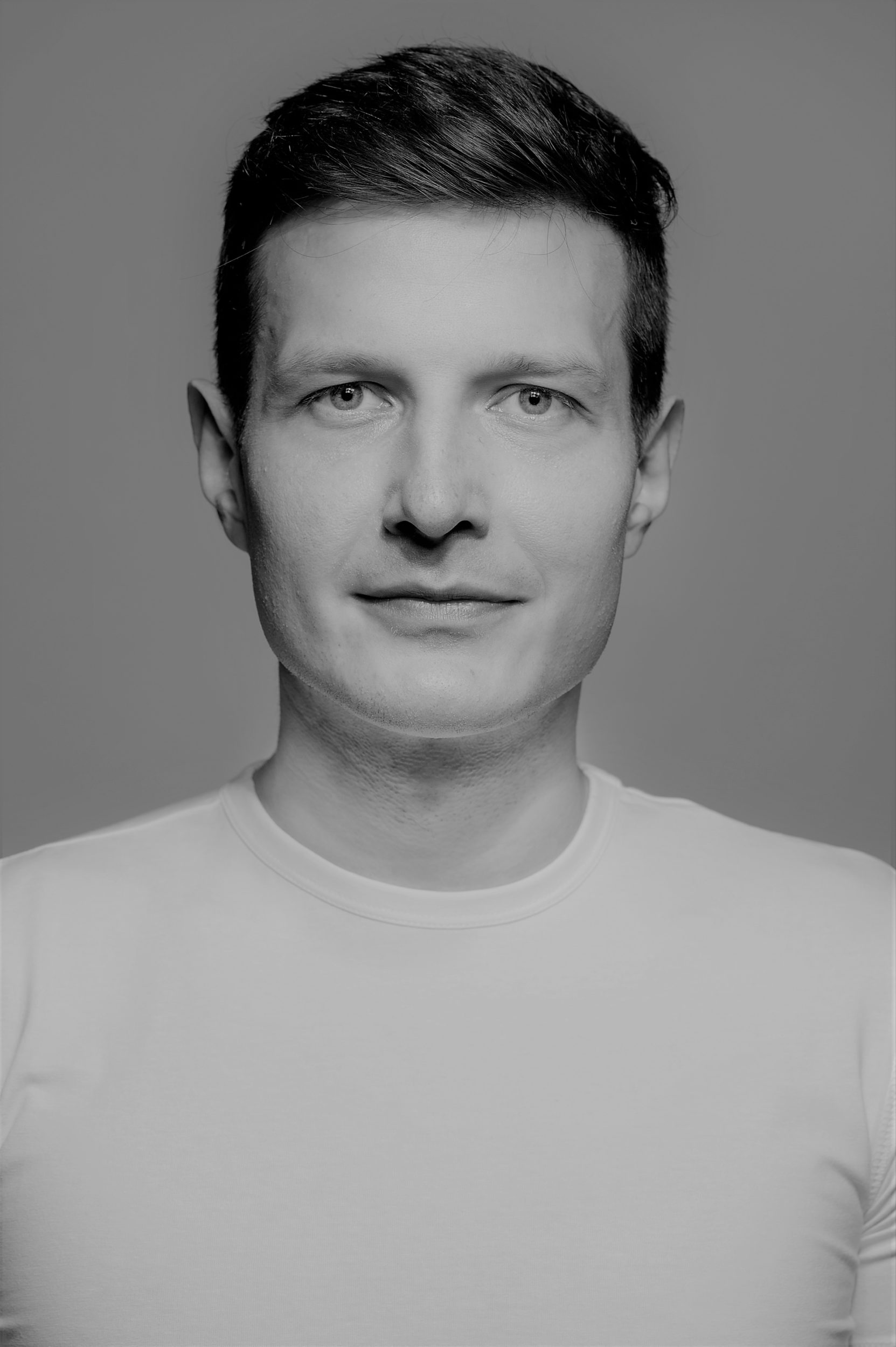 Cezary Jabłoński aktor. Portret mężczyzny, krótkie ciemne włosy, delikatny uśmiech, ubrany w jasny podkoszulek.