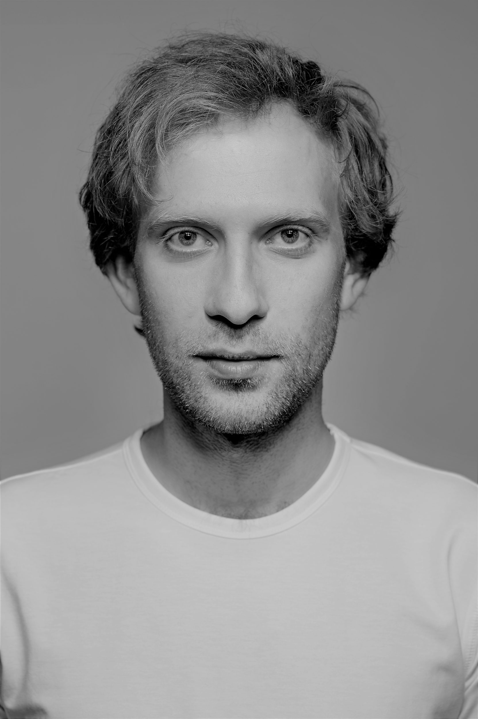 Mariusz Galilejczyk aktor. Portret mężczyzny, Falowane włosy do uszu, pogodna twarz, ubrany w biały podkoszulek.