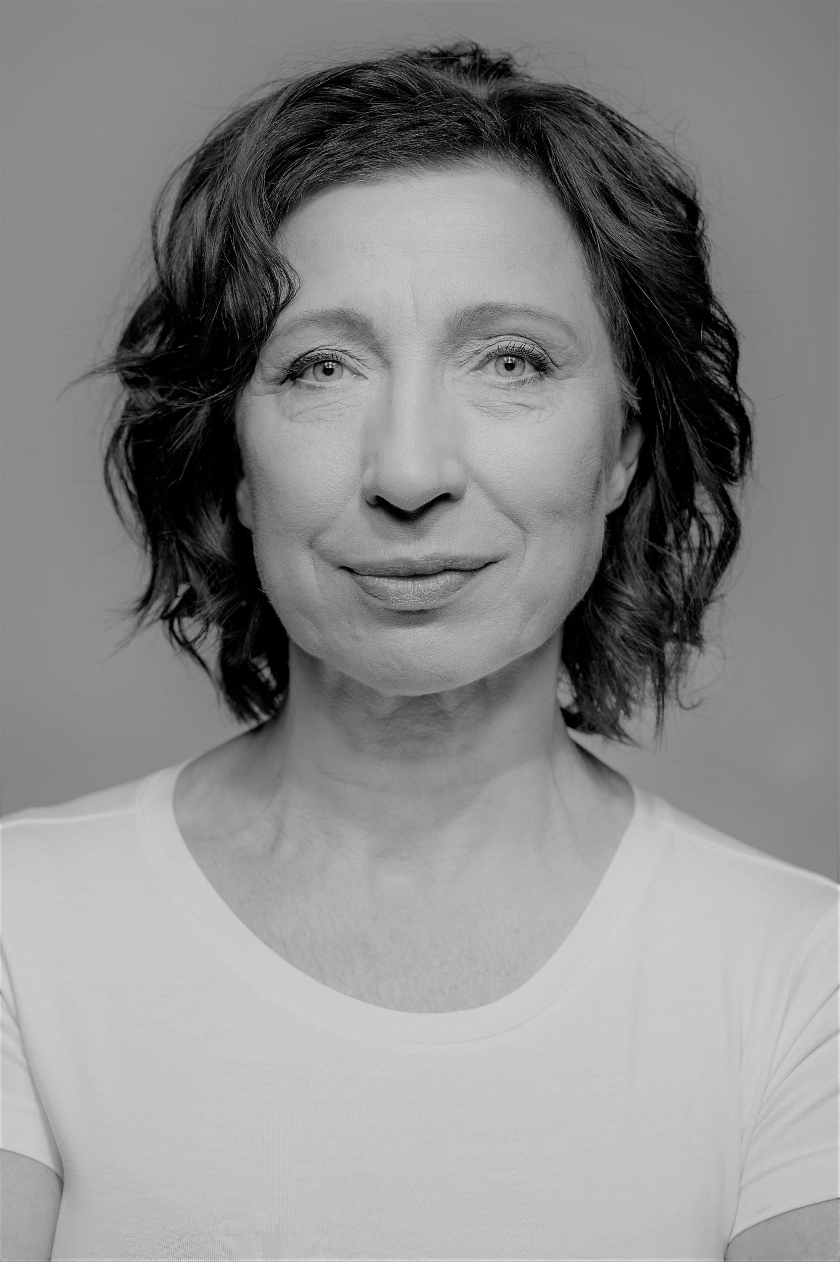 Jolanta Olszewska, portret. Kobieta, ciemne, falowane włosy nad ramiona, Ubrana w białą bluzkę.