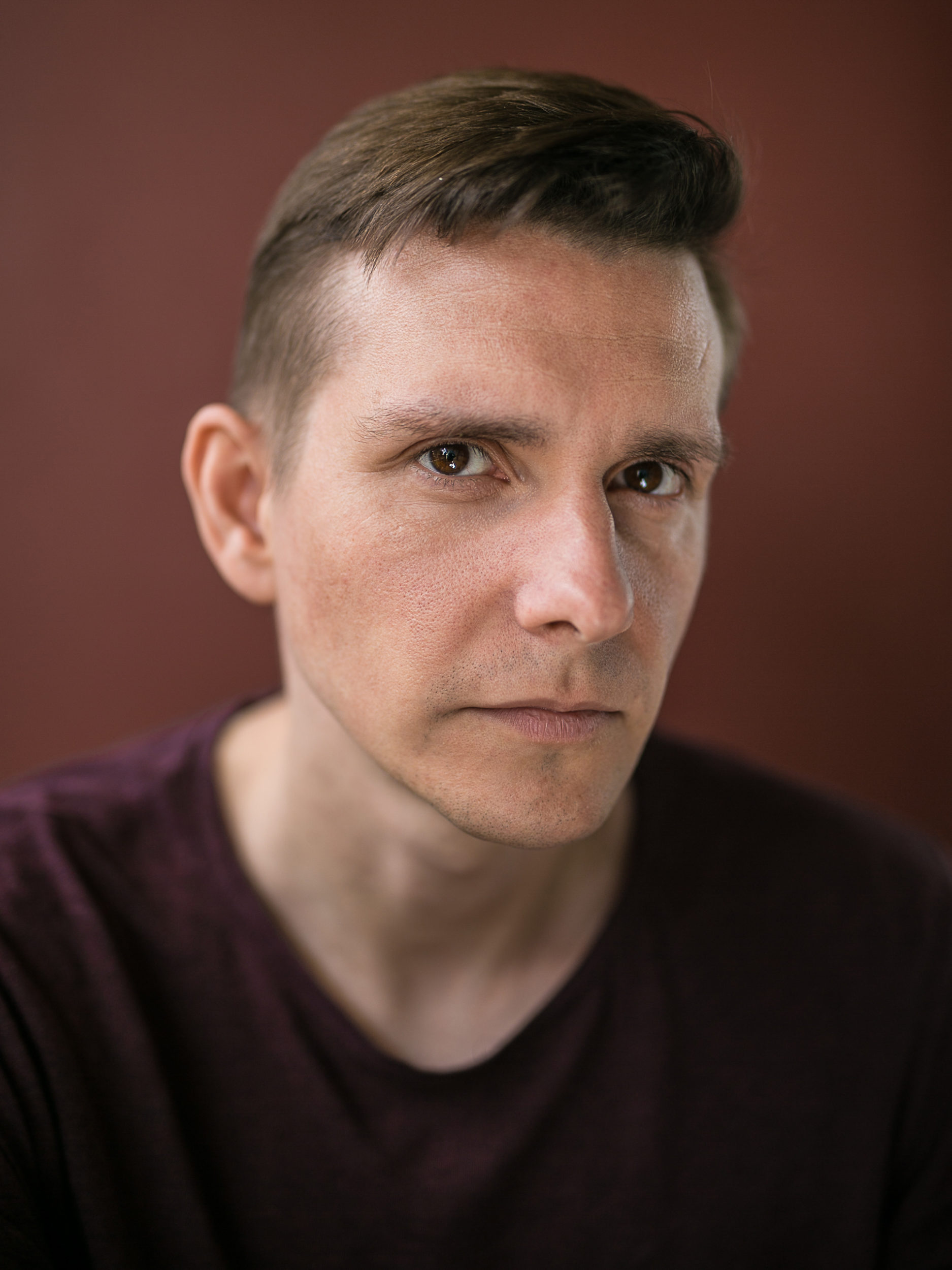 Paweł Majchrowski, aktor. Portret młodego mężczyzny na brązowym tle, Spokojna twarz, krótkie włosy, postawiona grzywka. Ubrany w ciemną bluzkę z dekoltem w serek.