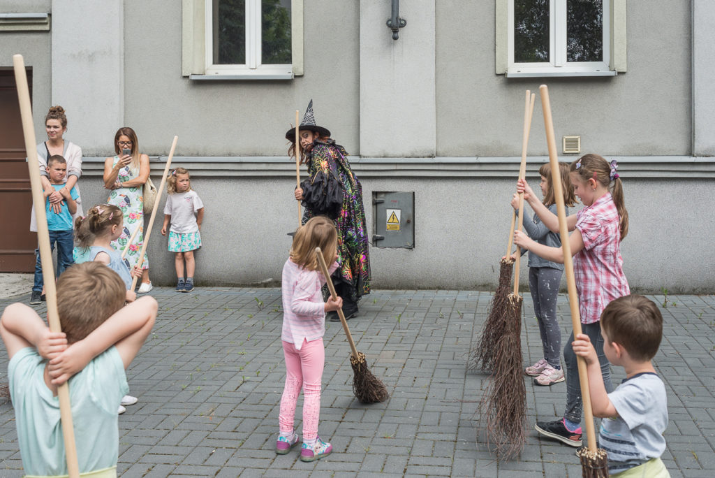 Dzieci z miotłami naśladują ruchy kobiety w kostiumie czarownicy.