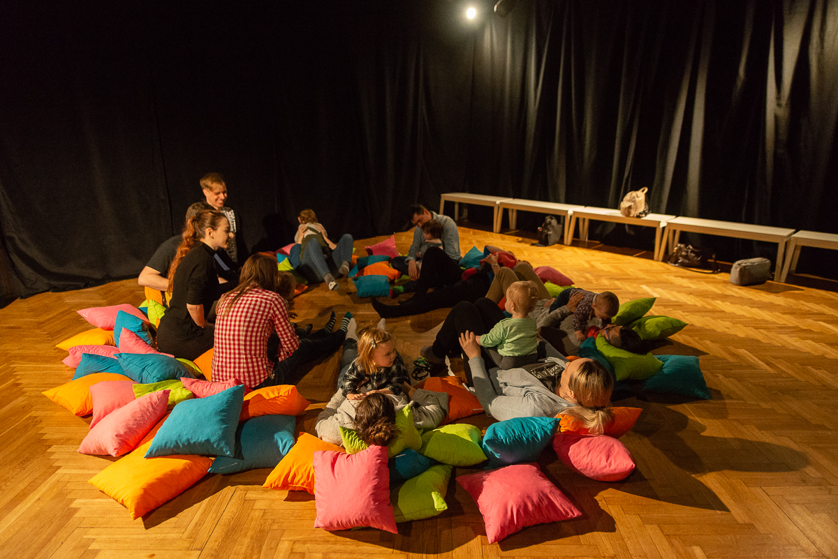 Dzieci i rodzice siedzą na podłodze na kolorowych poduszkach podczas warsztatów teatralnych.
