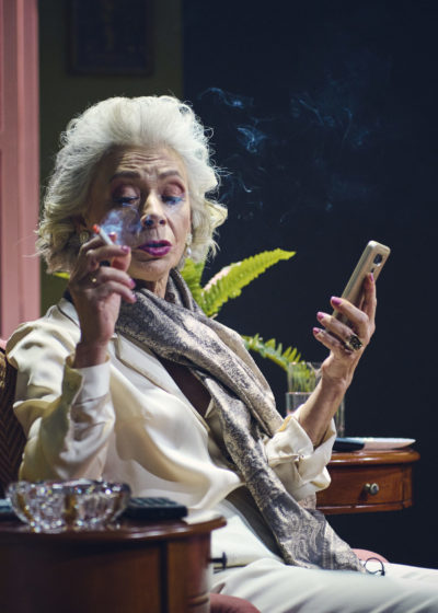 Starsza, elegancka kobieta siedzi w fotelu. W dłoni trzyma telefon komórkowy. Pali papierosa. W tle widać meble.