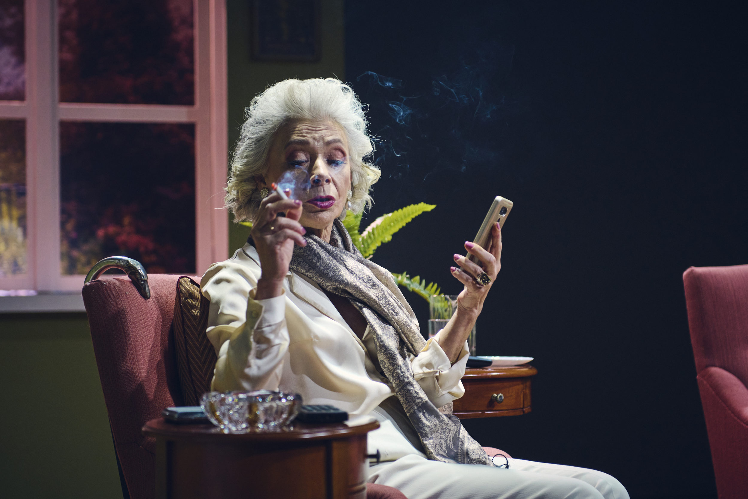 Starsza, elegancka kobieta siedzi w fotelu. W dłoni trzyma telefon komórkowy. Pali papierosa. W tle widać meble.