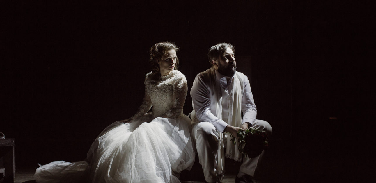 Na pustej scenie siedzi para aktorów. Kobieta i mężczyzna ubrani są na biało, rozmawiają. Mężczyzna trzyma bukiet róż.