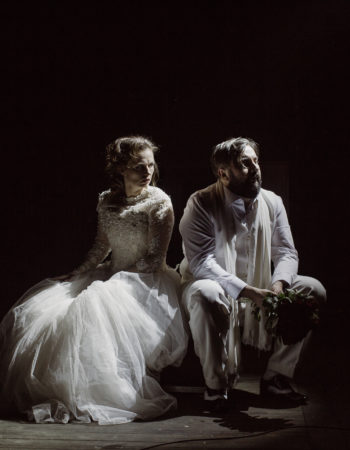 Na pustej scenie siedzi para aktorów. Kobieta i mężczyzna ubrani są na biało, rozmawiają. Mężczyzna trzyma bukiet róż.