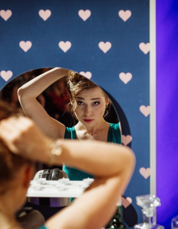 Elegancka kobieta siedzi tyłem. Jej twarz odbija się w lustrze. Kobieta poprawia sobie fryzurę.