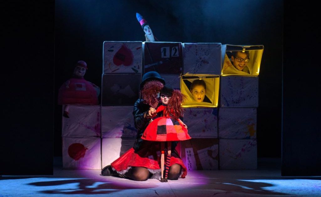 Aktorka trzyma lalkę czarownicy. W tle widać dekoracje z pudełek i twarze aktorów.