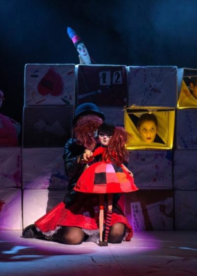 Aktorka trzyma lalkę czarownicy. W tle widać dekoracje z pudełek i twarze aktorów.