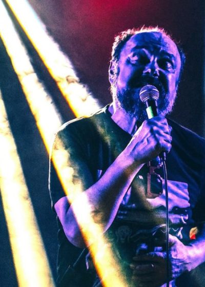 wokalista śpiewający do mikrofonu otulony światłami scenicznymi