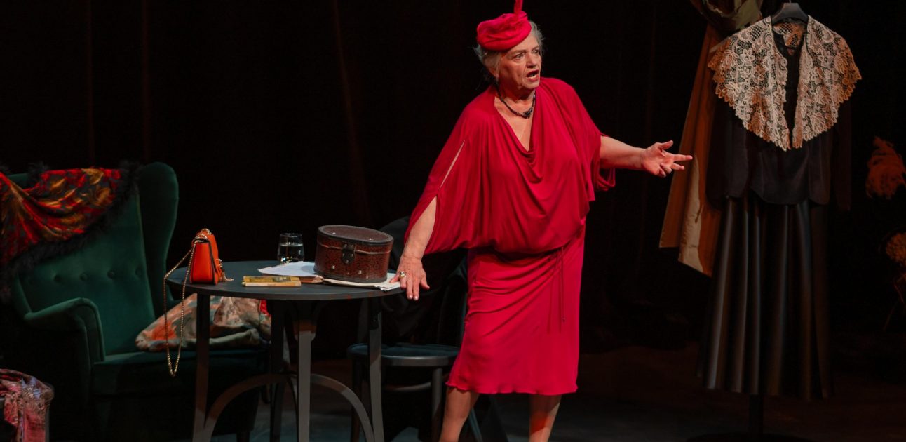 Anna Seniuk w czerwonej sukience i kapeluszu stoi oparta o stolik pośród starych mebli