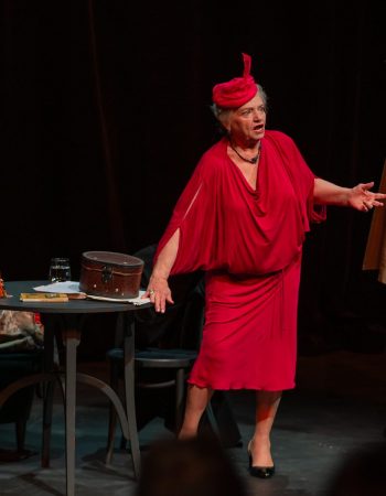 Anna Seniuk w czerwonej sukience i kapeluszu stoi oparta o stolik pośród starych mebli