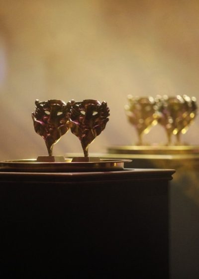 Na pierwszym planie dwie figurki złotych masek nagród konkursowych stojące na złotej tacy. Na drugim planie kolejne figurki.